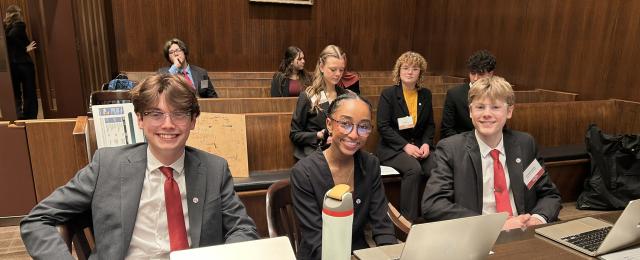 Student participating in the mock trial competition. Students sitting at an attorney table with other students sitting in spectator seats.